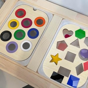 May include: Wooden educational toy with two sections. One side features colourful circles, the other has various shapes. The toy is set in a light-coloured wooden frame, designed for early learning and shape recognition.