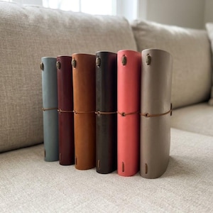 Puede incluir: Colección de siete diarios de cuero en varios colores, incluyendo azul, burdeos, naranja, marrón, coral y topo. Cada diario es cilíndrico, asegurado con un cordón de cuero y tiene un cierre decorativo. Los diarios están colocados sobre un sofá de tonos neutros.