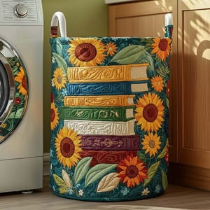 May include: A cylindrical laundry hamper with a dark teal background, adorned with embroidered sunflowers and a stack of colorful books. The hamper has white handles and is next to a washing machine.