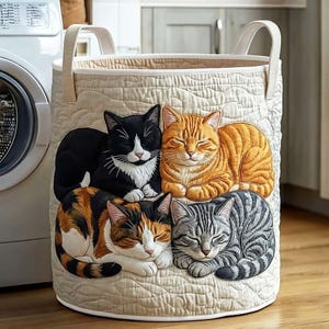 May include: A beige quilted laundry hamper featuring five sleeping cat appliqués in various colors: black and white, orange tabby, calico, and gray tabby. The hamper has two beige handles and a cream-colored interior.