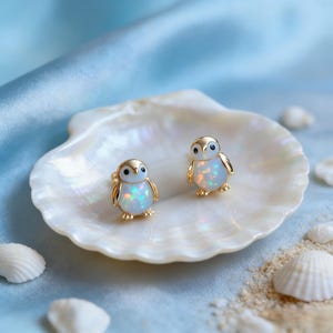 May include: A pair of gold-colored penguin stud earrings with opal-like bodies. The earrings are displayed on a white seashell, set against a backdrop of blue fabric and sand.