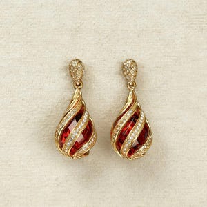 May include: A pair of gold-toned drop earrings with a teardrop design. Each earring features a vibrant red center stone, surrounded by a spiral of clear crystals and gold-colored metal. The top portion has a textured, diamond-like pattern.