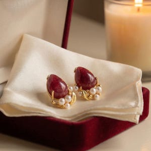 May include: A pair of ruby-colored gemstone earrings with gold accents and small pearl embellishments. The earrings are displayed on a cream-colored satin cloth inside a burgundy velvet box, suggesting a jewelry presentation.