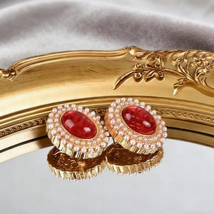May include: A pair of gold-toned stud earrings. Each earring features a large, oval, red gemstone surrounded by small, white pearls. The earrings are displayed on a reflective surface.