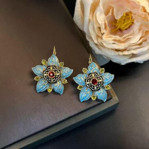 Könnte beinhalten: Ein Paar blütenförmige Ohrringe. Jeder Ohrring hat ein hellblaues Emaille-Blumendesign mit goldfarbenen Akzenten. Die Mitte jeder Blüte hat einen roten Edelstein, der von schwarzem und goldenem Filigran umgeben ist. Kleine grüne Edelsteine sind um die Blüte herum platziert.