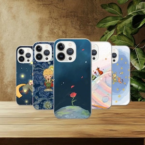 May include: A collection of five phone cases with colorful illustrations. The cases feature designs of a rose, a fox, a boy, and stars against a dark blue background. The cases are displayed on a wooden surface.