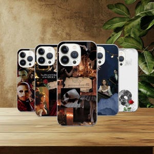 May include: A collection of five phone cases featuring designs inspired by "The Phantom of the Opera." The cases display images of the masked character, scenes from the musical, and the title text. The cases are on a wooden surface.