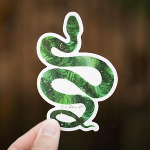 Op de afbeelding: Een groene slangsticker met een witte rand. Het slangontwerp heeft een marmereffect met tinten groen en goud. De sticker wordt door een hand vastgehouden. De handtekening van de kunstenaar, "Sarah Ribera Art", is zichtbaar.