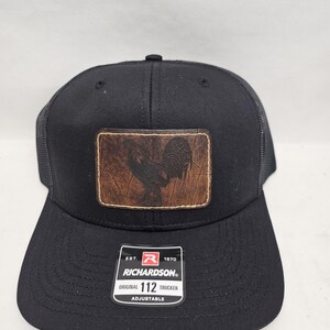 May include: Black trucker hat with a brown leather patch featuring a rooster design. The hat has a mesh back and a curved brim. The patch is rectangular and has a distressed look. The hat has the text "RICHARDSON" and "ORIGINAL 112 TRUCKER".