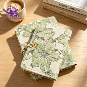 May include: Two rectangular notebooks with a cream-colored fabric cover featuring a green floral pattern. The notebooks are secured with a light-colored cord wrapped around two small, round, light brown wooden buttons on the front cover.