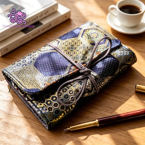 Op de afbeelding: Een rechthoekige stoffen etui met een donkerblauw en goudkleurig geometrisch patroon van zeshoekige motieven. De etui wordt gesloten met een dun, lichtbruin leren koord dat in een strik is geknoopt. Een vulpen van donker hout en goud ligt ernaast.