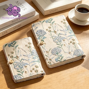 May include: Two cream-colored notebooks with textured covers featuring an embroidered floral pattern in shades of blue, green, and gold. Each notebook is secured with a small wooden button and an elastic loop closure on the front cover.