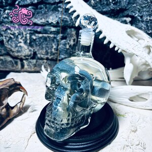 May include: A clear glass skull-shaped decanter with a crystal stopper. The decanter is filled with a blue liquid and sits on a black base. The background includes a skull and other bone fragments.