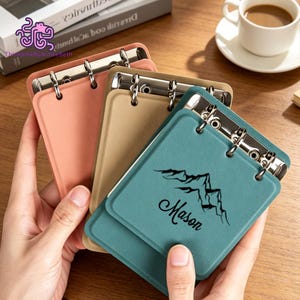 May include: Three small, rectangular, ring-bound notebooks in coral, tan, and teal. The teal notebook features a black mountain graphic and the name Mason printed in script. Each notebook has a silver metal ring mechanism at the top edge.