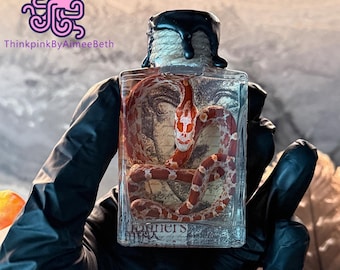 Op de afbeelding: Een heldere, rechthoekige glazen fles met een rood-witte slang erin. De slang heeft een schedelvormige markering op zijn kop. De fles is afgesloten met een met touw omwikkelde stopper en zwarte was. De tekst "Runners" is zichtbaar.