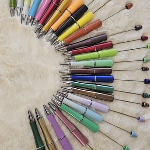 Op de afbeelding: Een verzameling kleurrijke pennen gerangschikt in een halve cirkel. De pennen hebben metallic zilveren accenten, met een verscheidenheid aan barrelkleuren, waaronder groen, geel, roze en blauw. Elke pen heeft een decoratieve top in bijpassende kleur.