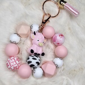 Op de afbeelding: Roze sleutelhanger met koeienmotief met een goudkleurige sluiting en kwastje. De sleutelhanger heeft een roze koeienbedel, roze, witte en patroon kralen. Een schattig en leuk accessoire.