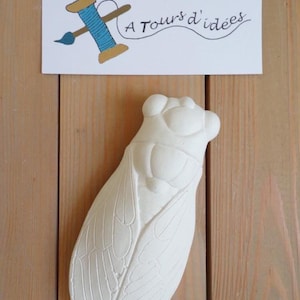 Peut inclure: Un objet décoratif blanc en forme de cigale avec des motifs d'ailes détaillés. L'image comprend une enseigne avec une bobine de fil et un pinceau, et le texte "A Tours d'idées". Le fond est une surface en bois.