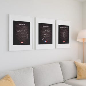 May include: Three framed prints on a wall. Each print features a map outline with text above and a flag at the bottom. The prints are labeled "Sisteron", "Cap Corse", and "Monaco Circuit". The prints are framed in white and hang above a gray sofa.