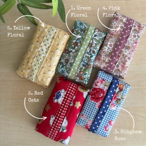 May include: Five rectangular fabric tissue holders featuring distinct patterns: yellow floral, green floral, pink floral, red with cat illustrations and gingham, and blue gingham with rose prints. Each holder has a central fabric strip.
