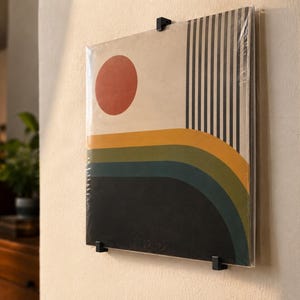 Könnte beinhalten: Eine quadratische Schallplattenhülle, die mit drei schwarzen Metallclips an der Wand befestigt ist. Das Cover zeigt ein grafisches Design mit einem großen orangefarbenen Kreis, vertikalen schwarzen Streifen und horizontalen gelben, grünen und dunkelblauen Bändern.