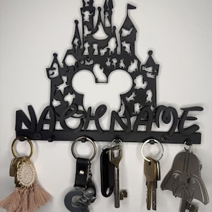 Könnte beinhalten: Ein schwarzer Wand-Schlüsselhalter mit einer Schloss-Silhouette, integrierten Figuren-Ausschnitten und einer zentralen Mickey-Maus-Kopfform. Das Wort NACHNAME ist in erhabenen Buchstaben über fünf Haken mit verschiedenen Schlüsseln angebracht.