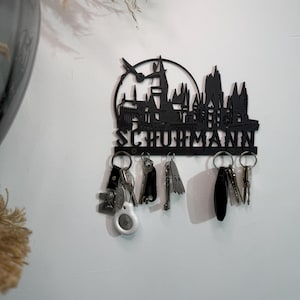 Könnte beinhalten: Ein schwarzer Wand-Schlüsselhalter mit der Silhouette einer Burg und einer fliegenden Figur darüber. Der Name SCHUHMANN ist unten in Großbuchstaben ausgeschnitten. Fünf Metallhaken halten verschiedene Schlüssel und einen weißen Tracker.