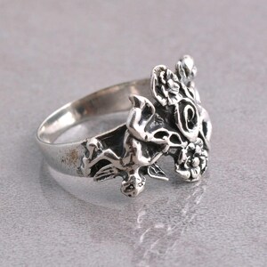 May include: A silver ring featuring a detailed floral and cherub design. The ring has a textured finish and intricate detailing, showcasing a classic, vintage aesthetic. The ring is set against a neutral background.