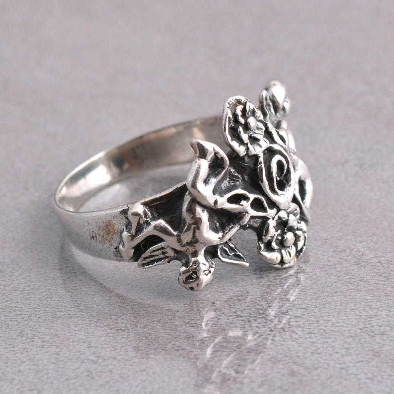 May include: A silver ring featuring a detailed floral and cherub design. The ring has a textured finish and intricate detailing, showcasing a classic, vintage aesthetic. The ring is set against a neutral background.