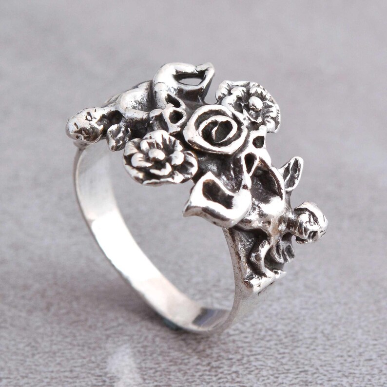 May include: A silver ring featuring a floral design with detailed flowers and leaves. The ring has a polished finish and is set against a gray background. The ring's design includes a rose and other floral elements.
