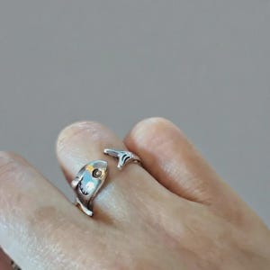 Puede incluir: Un anillo plateado con forma de pez. El anillo presenta un diseño detallado de pez, con la cabeza y la cola formando los extremos del anillo. El pez tiene un ojo y aletas. El anillo se lleva en un dedo.