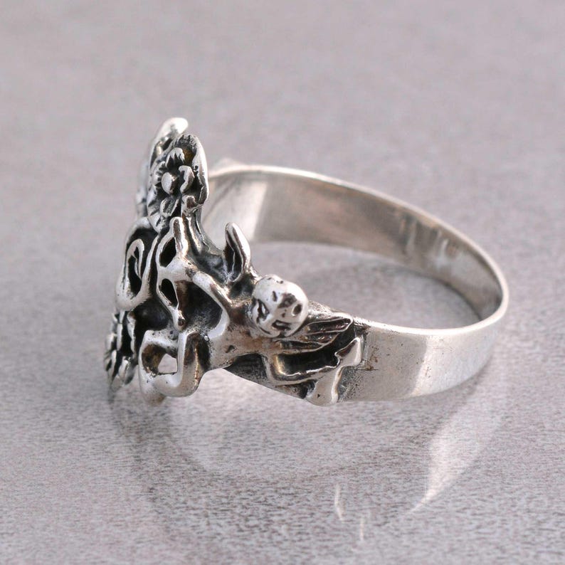 May include: A silver ring featuring a detailed cherub design. The ring has a wide band and a sculpted cherub with wings and floral accents. The ring is made of a shiny, metallic material and is set against a neutral background.