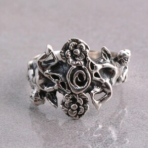May include: A silver-toned ring with an intricate floral design. The ring features multiple flowers and swirling patterns, creating a detailed and ornate appearance. The metal has a slightly oxidized finish, enhancing the details.