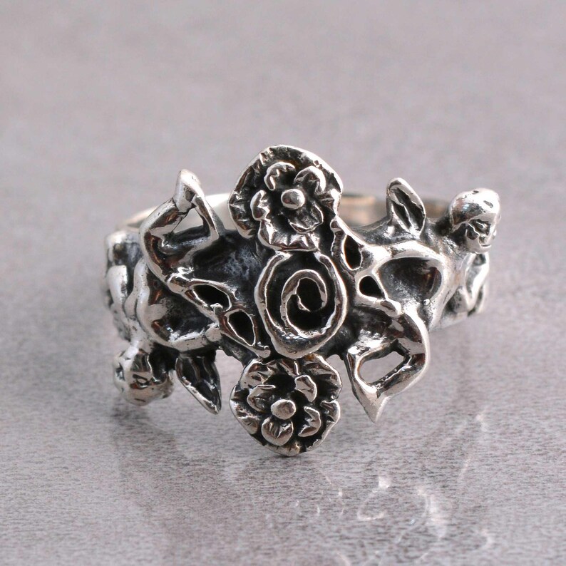 May include: A silver-toned ring with an intricate floral design. The ring features multiple flowers and swirling patterns, creating a detailed and ornate appearance. The metal has a slightly oxidized finish, enhancing the details.