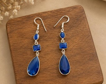 May include: A pair of silver dangle earrings featuring a vertical arrangement of three blue stones. Each earring consists of a small oval stone, a square stone, and a large teardrop-shaped stone suspended from a silver hook wire.