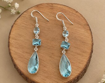 May include: A pair of silver drop earrings featuring three light blue faceted stones in oval, rectangular, and teardrop shapes. The earrings hang from silver hooks and are arranged in a vertical line with the largest stone at the bottom.