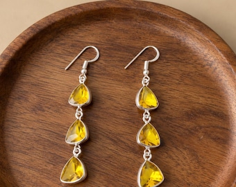May include: A pair of silver drop earrings, each featuring three faceted, teardrop-shaped yellow stones arranged vertically. The earrings are suspended from silver hooks and have a polished metallic finish, showcasing a clear, bright yellow hue.