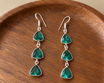 May include: A pair of silver drop earrings, each featuring three faceted, triangular emerald green stones arranged vertically. The earrings are suspended from silver hooks and have a polished metal setting that secures each stone.
