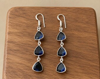 May include: A pair of silver drop earrings, each featuring three faceted, triangular dark blue stones arranged vertically. The earrings are suspended from silver hook wires, showcasing a clean, metallic finish and deep blue hues.