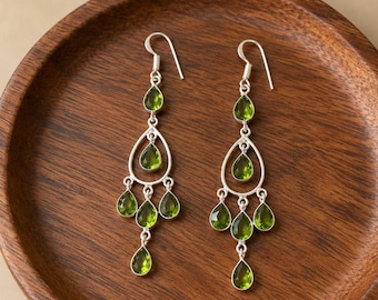 May include: A pair of silver chandelier earrings featuring multiple pear-shaped green peridot gemstones. Each earring has a hook wire and a tiered design with one stone at the top, one in the center of a teardrop frame, and three stones dangling below.