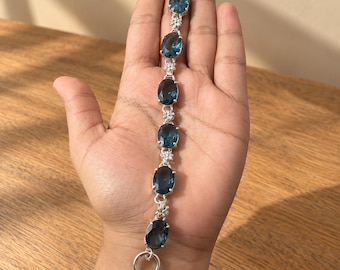 May include: A silver bracelet featuring six oval-cut dark blue gemstones arranged in a vertical line. Each gemstone is separated by a small, intricate silver floral-shaped link. The bracelet is finished with a silver toggle clasp closure.