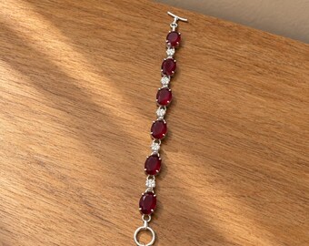 May include: A silver-toned bracelet featuring a series of alternating oval-shaped deep red stones and small clear round stones. The bracelet is finished with a toggle clasp consisting of a T-bar and two circular rings at one end.