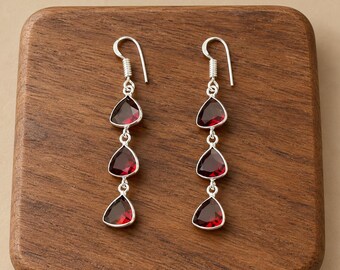 May include: A pair of drop earrings featuring three faceted, teardrop-shaped deep red stones set in silver-toned metal. Each earring hangs from a simple silver hook, with the stones arranged vertically in a graduated, linear design.
