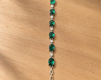 May include: A silver-toned bracelet featuring five oval-cut emerald green stones alternating with small, clear, round-cut stones. The bracelet includes a toggle clasp at one end and a circular link at the other, with a polished metal chain design.