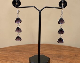 May include: A pair of dangle earrings featuring three faceted, triangular purple stones set in silver-toned metal. Each earring hangs from a silver-toned hook, with the stones arranged vertically in a column, connected by small silver-toned rings.