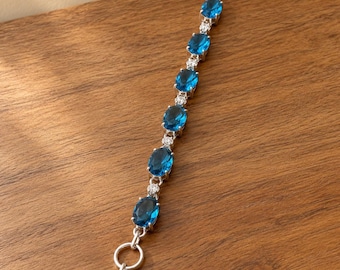 May include: A silver-toned bracelet featuring seven oval-cut blue gemstones arranged in a line, alternating with small clear round stones. The piece includes a toggle clasp closure with a circular ring and a T-bar connector at the end.