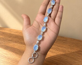 May include: A silver-toned bracelet featuring seven oval-shaped, translucent light blue stones arranged in a row. The stones are connected by small silver-toned metal links, and the bracelet ends with a circular clasp mechanism.