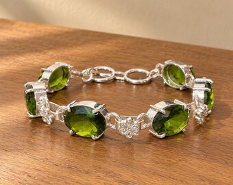 May include: A silver link bracelet featuring oval-shaped, translucent olive green gemstones set in prongs. The bracelet alternates between these green stones and small, six-petaled silver flower accents, finished with a circular link clasp.