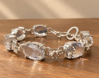May include: A silver-toned link bracelet featuring multiple oval-shaped clear faceted stones set in metal prongs. The bracelet includes small flower-shaped metal connectors between the stones and a circular toggle clasp closure.