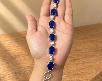 May include: A silver-toned bracelet featuring a series of six oval-shaped, deep blue faceted stones alternating with small, clear, round accent stones. The piece is finished with a circular toggle clasp at the end of the chain.